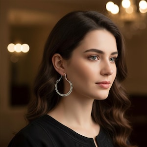 Puede incluir: Una mujer lleva pendientes de aro plateados con pequeños adornos redondos. Los pendientes son de diseño clásico y la mujer lleva una blusa negra. El fondo está borroso, con iluminación cálida.