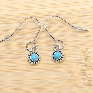 Puede incluir: Un par de pendientes plateados con un centro de piedra turquesa y un borde decorativo. Los pendientes tienen un cierre de gancho. Se muestran sobre una superficie de madera marrón claro.