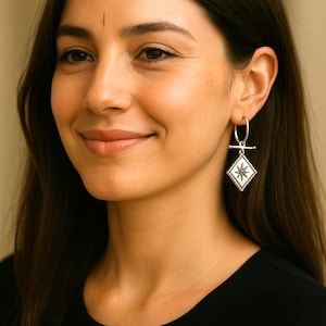 Puede incluir: Pendientes de aro plateados con un colgante geométrico en forma de diamante. El colgante presenta un diseño de estrella en el centro, rodeado por un borde punteado. La mujer de la imagen lleva una camiseta negra.