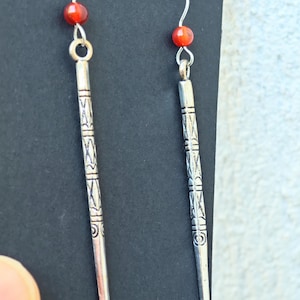 Puede incluir: Un par de pendientes plateados con una cuenta roja y un diseño largo grabado. Los pendientes tienen un cierre de gancho y se muestran sobre un fondo negro.