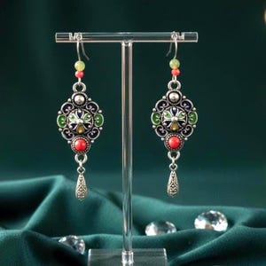 Puede incluir: Un par de pendientes plateados adornados con detalles de esmalte colorido. Cada pendiente presenta un diseño floral central en azul, verde y amarillo, acentuado con cuentas rojas y verdes. Los pendientes cuelgan de un gancho plateado y tienen un colgante en forma de lágrima.