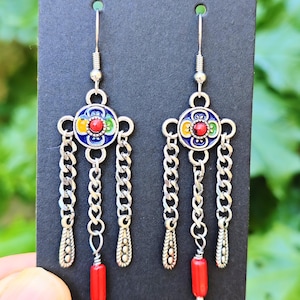 Puede incluir: Pendientes colgantes plateados con un diseño floral colorido y detalles de cadena. Cada pendiente presenta una piedra roja central rodeada de esmalte azul, amarillo y verde. Los pendientes tienen tres cadenas y una cuenta cilíndrica roja.