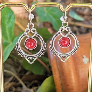 Puede incluir: Un par de pendientes plateados con una gema roja redonda en el centro. Los pendientes tienen un diseño calado ornamentado con un marco circular y detalles decorativos. Se exhiben sobre un fondo borroso de follaje verde.