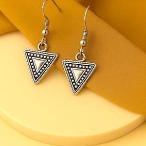 Puede incluir: Pendientes triangulares plateados con borde de cuentas e interior punteado. Los pendientes cuelgan de un gancho curvo y una bolita. Los pendientes se exhiben sobre una superficie amarilla.
