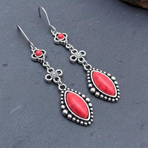 Puede incluir: Pendientes colgantes plateados con piedras rojas de forma ovalada. Los pendientes presentan un diseño floral y pequeños detalles redondos alrededor de las piedras rojas. Los ganchos son plateados.