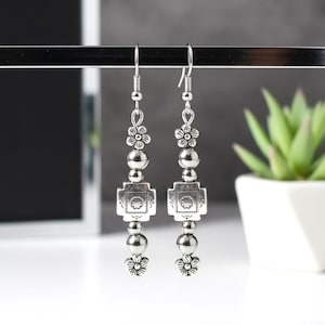 Puede incluir: Pendientes colgantes plateados. Cada pendiente presenta un diseño floral en la parte superior e inferior, con una pieza central cuadrada en forma de cruz. Los pendientes están compuestos por cuentas de varios tamaños y cuelgan de un gancho.