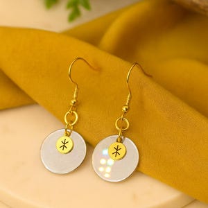 Puede incluir: Un par de pendientes colgantes dorados. Cada pendiente presenta un dije circular iridiscente con un dije de asterisco dorado. Los pendientes cuelgan de ganchos dorados. El fondo es una tela de color amarillo mostaza.