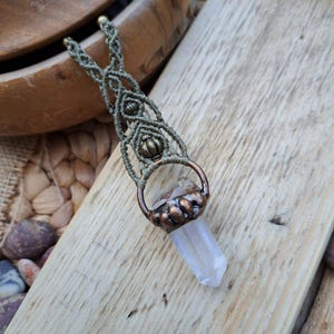 May include: A clear quartz crystal point pendant with a copper wire wrap and green macrame cord. The pendant is hanging from a wooden surface.