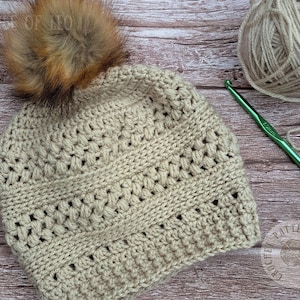 May include: A beige crocheted beanie with a brown faux fur pom-pom. The hat features a textured stitch pattern and is displayed with a ball of yarn and a crochet hook. The hat is handmade and suitable for cold weather.
