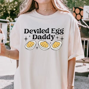 Puede incluir: Camiseta color crema con el texto "Deviled Egg Daddy" sobre un gráfico de tres huevos rellenos. La camiseta es de corte holgado y tiene mangas cortas. El gráfico es en negro y amarillo.