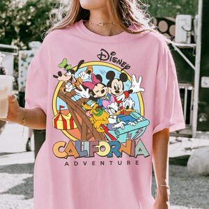 May include: A light pink t-shirt featuring a Disney graphic with Mickey Mouse, Minnie Mouse, Donald Duck, Goofy, and other characters on a roller coaster. The text "CALIFORNIA ADVENTURE" is below the graphic.