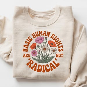 以下が含まれることがあります： オレンジ、ピンク、白の花のイラストが描かれた、"BASIC HUMAN RIGHTS are NOT 'RADICAL'"というテキストが円形にデザインされた、クリーム色のスウェットシャツ。長袖は折りたたまれています。