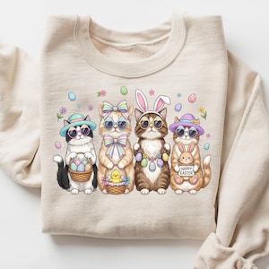 May include: Beige sweatshirt with an Easter-themed graphic. The design shows four cats in sunglasses, hats, and bunny ears, each with Easter baskets or signs. The graphic includes pastel eggs and floral details. The sweatshirt is folded.