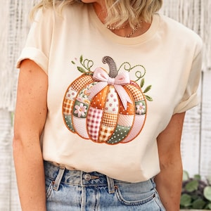 以下が含まれることがあります： パッチワークのかぼちゃのデザインとピンクのリボンが付いた、クリーム色のTシャツ。かぼちゃは、オレンジ、ピンク、緑、白のさまざまな柄の生地で構成されています。このシャツは、青いジーンズと組み合わせています。