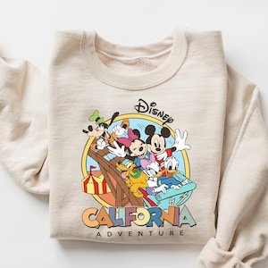 May include: Beige crewneck sweatshirt featuring a colorful graphic of Mickey Mouse, Minnie Mouse, Donald Duck, Goofy, and Pluto. The graphic is set within a yellow circle and includes the text "Disney California Adventure".