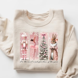 Puede incluir: Sudadera color crema con un diseño festivo. El gráfico incluye un cascanueces rosa, una bailarina, un árbol de Navidad y un ratón con corona. El texto dice: "It's the most wonderful time of the year".