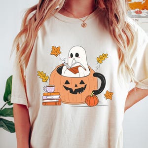 May include: Beige t-shirt featuring a Halloween-themed graphic. The design includes a ghost reading a book inside a pumpkin mug, surrounded by autumn leaves and a small pumpkin. A stack of books and a teacup are also present.
