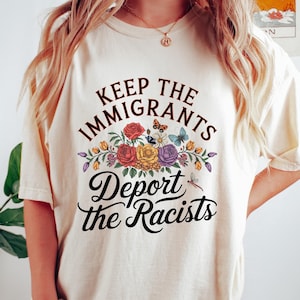 May include: A cream-coloured t-shirt featuring the slogan "Keep the Immigrants, Deport the Racists" in black script. The text is above a floral design with red, yellow, and purple flowers, and butterflies. The shirt is a casual style.