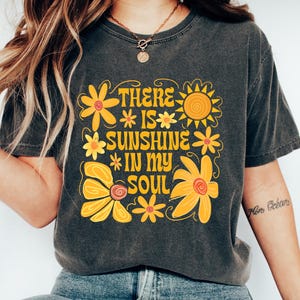 May include: Charcoal grey t-shirt featuring a retro floral design and the text "THERE IS SUNSHINE IN MY SOUL" in yellow. The shirt is worn with blue jeans and white trainers. The style has a positive, vintage aesthetic.