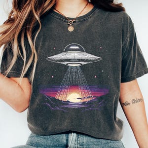 Op de afbeelding: Donkergrijs T-shirt met een gepixeld grafisch ontwerp van een UFO die licht schijnt op een zonsondergang. Het ontwerp omvat een gedetailleerd ruimteschip en een levendige zonsondergang met paarse en oranje tinten. Het is een casual kledingstuk.