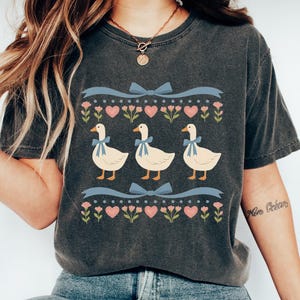 May include: Dark gray t-shirt featuring a whimsical design of three geese with blue bows, set against a backdrop of pink hearts and flowers. The shirt has a vintage, washed look, and is part of a clothing collection.