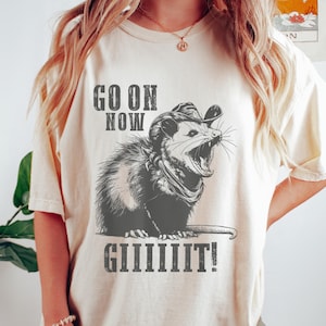 Puede incluir: Camiseta color crema con una ilustración en blanco y negro de una zarigüeya con sombrero de vaquero y pañuelo. El texto "GO ON NOW GIIIIIIIT!" está impreso en negrita, con un efecto desgastado. La camiseta tiene mangas cortas.