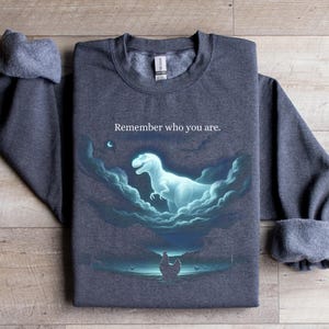 May include: Dark gray sweatshirt with a graphic of a glowing dinosaur in clouds, with the text "Remember who you are." The design is in shades of blue and white. The sweatshirt has a crew neck and long sleeves.