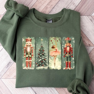 Puede incluir: Sudadera verde con un diseño navideño vintage. El diseño incluye dos cascanueces, un árbol de Navidad y una bailarina, enmarcados en rectángulos sobre un fondo nevado. La sudadera está hecha de un material suave.