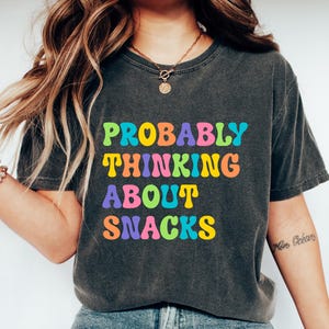 Pode incluir: T-shirt cinza escuro com o texto "PROBABLY THINKING ABOUT SNACKS" em letras coloridas de estilo retro. A camisa é combinada com jeans azuis e tênis brancos. Um traje casual e confortável.