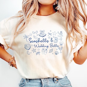 May include: Cream-colored t-shirt with the text "Seashells & Wedding Bells" in blue, surrounded by nautical and wedding-themed illustrations. The shirt is paired with light blue jeans.