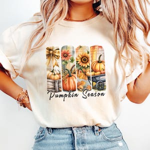 May include: Cream-colored t-shirt with a colorful graphic featuring pumpkins, sunflowers, and the text "Pumpkin Season." The design includes a wooden crate, a wheelbarrow, and fall foliage. The shirt is paired with light blue jeans.