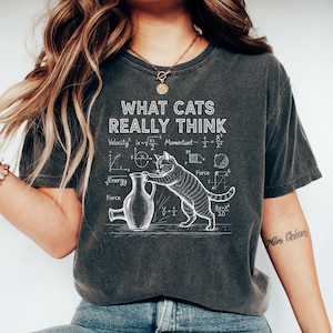 Può includere: T-shirt grigio scuro con la scritta "WHAT CATS REALLY THINK" e una grafica di un gatto che interagisce con un vaso, circondato da equazioni matematiche. Stile casual e confortevole.
