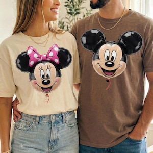 Peut inclure: Un t-shirt cr&egrave;me avec un motif de ballon Minnie Mouse portant un n&oelig;ud rose &agrave; pois et un t-shirt marron avec un motif de ballon Mickey Mouse. Les deux t-shirts ont des manches courtes et un col rond, mettant en valeur des personnages embl&eacute;matiques.