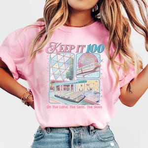 May include: Pink t-shirt with a graphic design featuring the text "KEEP IT 100" and illustrations of a theme park. The design includes images of a monorail, a boat ride, and a geodesic sphere. The text "On The Land, The Seas, The Skies" is also included.