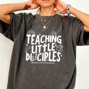 Könnte beinhalten: Dunkelgraues T-Shirt mit dem weißen Text "TEACHING LITTLE DISCIPLES". Das Design umfasst einen Regenbogen, eine Sonne, einen Blitz, eine Krone und eine Taube. Das T-Shirt hat kurze Ärmel und eine lockere Passform.