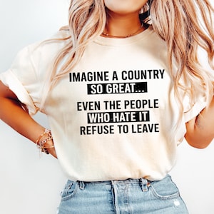 May include: Cream-colored t-shirt with the text "IMAGINE A COUNTRY SO GREAT... EVEN THE PEOPLE WHO HATE IT REFUSE TO LEAVE" in black. The shirt is paired with light blue denim jeans.