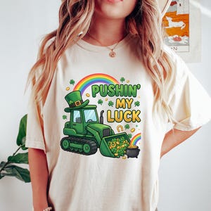Puede incluir: Camiseta color crema con un diseño del Día de San Patricio. El gráfico incluye una excavadora verde con un sombrero de duende, un arcoíris, tréboles y el texto "Pushin' My Luck". También se ve una olla de oro.