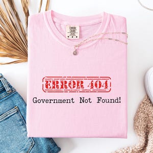 以下が含まれることがあります： 「ERROR 404 Government Not Found!」の文字が赤いスタンプ風のデザインでプリントされた、薄いピンク色のTシャツ。白い表面に折りたたまれており、青いジーンズ、白いスニーカー、ネックレスが近くに置かれています。