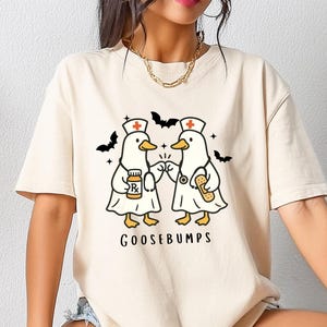 May include: Cream-colored t-shirt featuring a cartoon design of two ducks dressed as nurses, with bats and the word "GOOSEBUMPS". The ducks are holding medical items. The shirt is a casual, short-sleeved style.