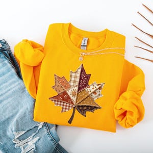 May include: A bright yellow crewneck sweatshirt featuring a large, central graphic of a maple leaf composed of various patchwork fabric patterns in shades of brown, cream, and burgundy, paired with light blue distressed denim jeans.
