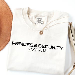 Może przedstawiać: Kremowa koszulka z czarnym napisem "PRINCESS SECURITY SINCE 2013". Widoczny jest również złoty naszyjnik z wisiorkiem i brązowy kapelusz. Koszulka wykonana jest z miękkiego materiału.