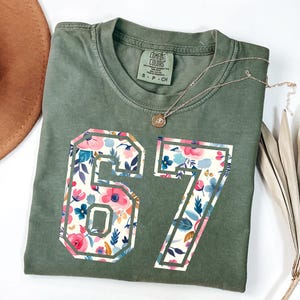 May include: Olive green t-shirt with the numbers 67 in a floral pattern. The numbers are outlined in a white border. A gold necklace with a pendant is on the shirt.