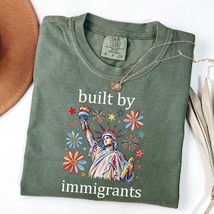 May include: An olive green t-shirt featuring a graphic of the Statue of Liberty surrounded by colorful flowers and stars. The text "built by immigrants" is printed in white above and below the graphic. The shirt has a crew neck and short sleeves.