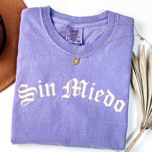 May include: A lavender Comfort Colors t-shirt with the phrase "Sin Miedo" in a cream-colored gothic font. A gold necklace with a pendant rests on the shirt. A brown hat and dried palm leaf are in the background.