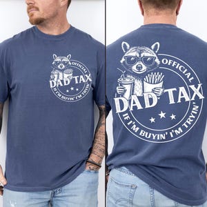 May include: A navy blue t-shirt featuring a raccoon wearing sunglasses, holding a drink and fries, with the text "DAD TAX" and "IF I'M BUYIN' I'M TRYIN" in a circular design. The shirt is a casual wear item.