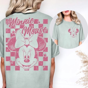 May include: A pale blue t-shirt with a pink Minnie Mouse design. The graphic shows Minnie's face, bow, and the words "Minnie Mouse" in a distressed style. The design is set against a pink and white chequered background. A smaller version of the graphic is on the pocket.