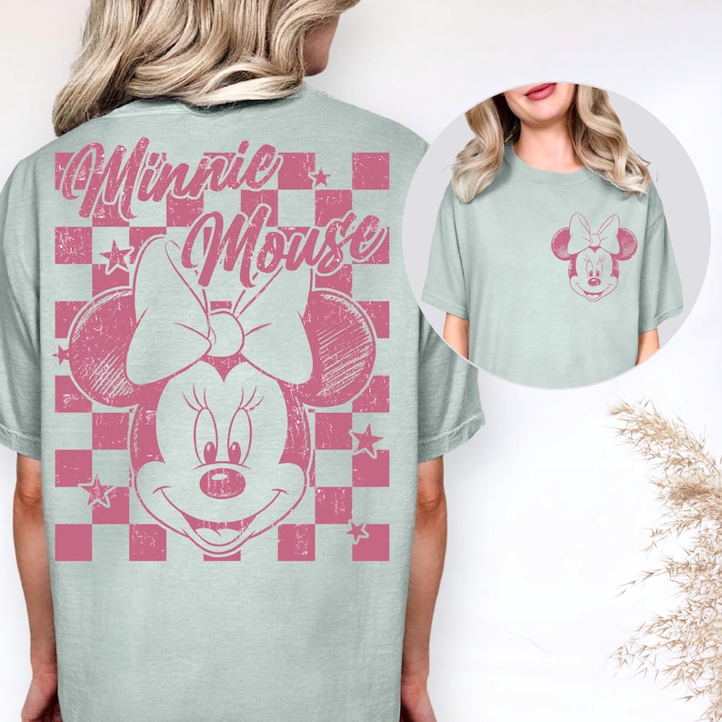 May include: A pale blue t-shirt with a pink Minnie Mouse design. The graphic shows Minnie's face, bow, and the words "Minnie Mouse" in a distressed style. The design is set against a pink and white chequered background. A smaller version of the graphic is on the pocket.