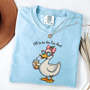 Puede incluir: Una camiseta azul claro con el gráfico de un pato blanco que lleva gafas de sol en forma de corazón y un lazo rosa, sosteniendo un vaso de zumo de naranja con una pajita. El texto superior dice, Off to be the Fun Aunt. Etiqueta Comfort Colors.