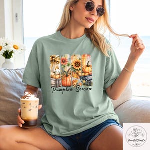 May include: Sage green t-shirt featuring a fall-themed graphic with pumpkins, sunflowers, and the text "Pumpkin Season." The design includes a wooden crate, a wheelbarrow, and autumn leaves. The shirt is a comfortable, casual style.