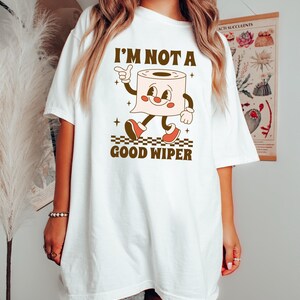 May include: White t-shirt with a retro cartoon toilet paper roll character. The character has a smiling face, arms, and legs, wearing red shoes. The text "I'M NOT A GOOD WIPER" is printed above and below the character in brown.